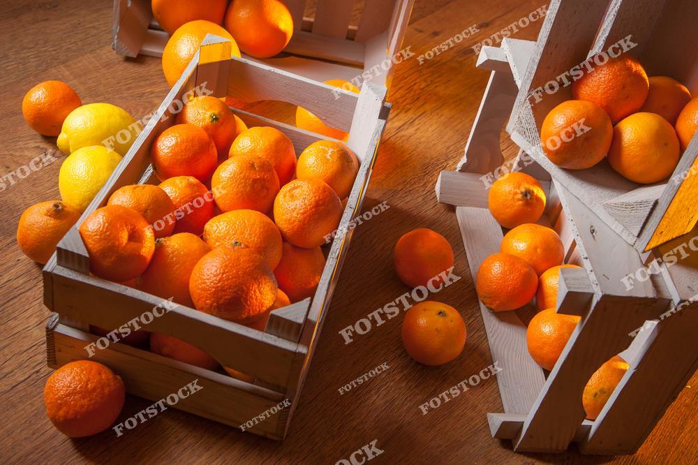 На фотографии изображены деревянные ящики, наполненные мандаринами и лимонами, расположенные на деревянном столе. Некоторые фрукты рассыпаны вокруг, что добавляет динамики композиции. Снимок передает ощущение натуральности, изобилия и зимнего настроения.